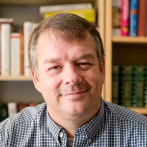 Matthew Coburn, a white man with short greying hair, smiling in front of a bookshelf