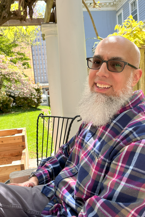 A photo of Omer Bajwa, a bald asian man with a long grey beard and glasses, outdoors, smiling in a plaid shirt.