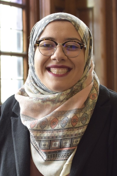 Leenah Safi, a light skinned woman in a dark jacket, glasses and patterned pastelle hijab, smiling at the camera.