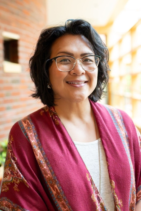 Pauline Little, an asian woman with shoulder length black curly hair and glasses, indoors, smiling towards the camera.