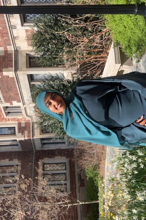 Afnan Shure, a brown skinned woman wearing a teal hijab, smiling towards the camera, surrounded by daffodils and collegiate gothic architecture.