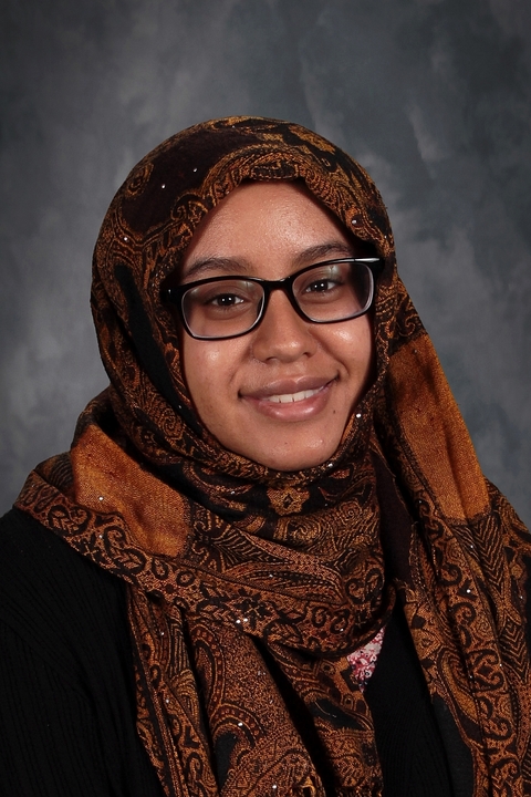 Sarah Mahmoud, a dark skinned woman wearing black rimmed glasses and a patterned orange hijab, in front of a grey backdrop