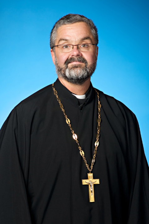 Father Voytovich, a white man with short grey hair and beard, in clerical dress and glasses in front of a blue backdrop