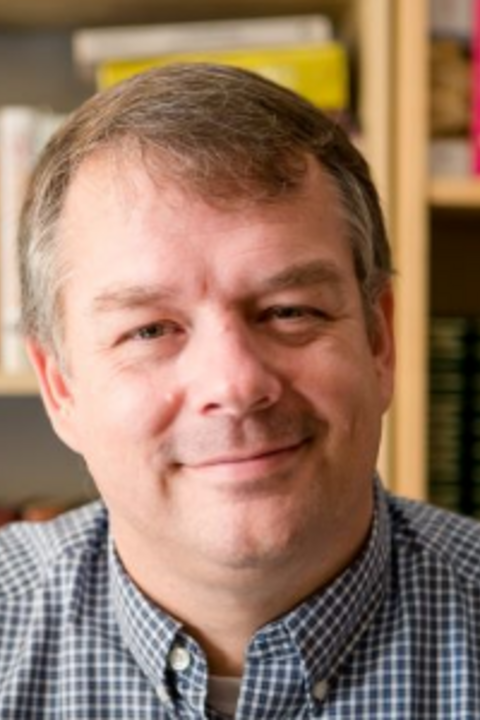 Matthew Coburn, a white man with short greying hair, smiling in front of a bookshelf
