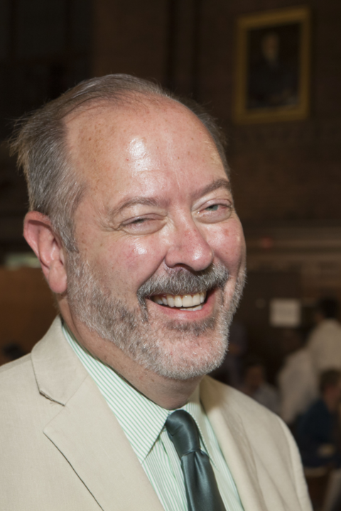 Ian Oliver, a bald white man with short grey hair and a grey beard, smiling towards the camera in a cream colored suit.