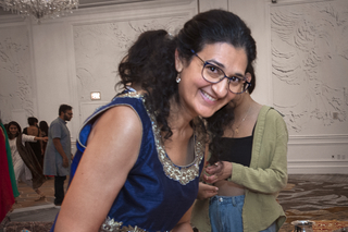 Asha Shipman, a South asian woman with shoulder length curly black hair and glasses, lighting diwali candles while wearing a blue dress.