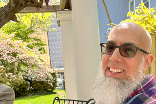 A photo of Omer Bajwa, a bald asian man with a long grey beard and glasses, outdoors, smiling in a plaid shirt.