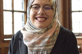 Leenah Safi, a light skinned woman in a dark jacket, glasses and patterned pastelle hijab, smiling at the camera.