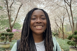 A photo of Erica Adarkwa, a Black woman with shoulder length dredlocks, smiling at the camera with blossoming trees in the background.