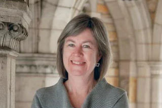 Sharon Kugler, a white woman with shoulder length grey hair, smiling in front of collegiate gothic architecture