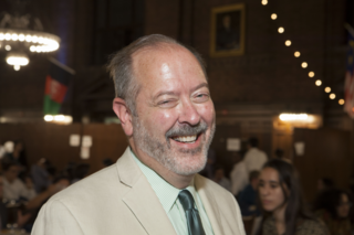 Ian Oliver, a bald white man with short grey hair and a grey beard, smiling towards the camera in a cream colored suit.